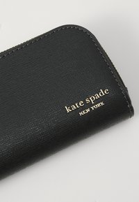 Portefeuille noir texturé avec le logo "kate spade NEW YORK" doré, doté d'une fermeture éclair et de coins arrondis. Matière en cuir à grain lisse.