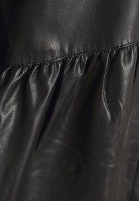 Schwarzes Leder mit glatter Textur und dezentem Glanz, mit gerafften Falten entlang des Saums. Die Nähte sind an den Kanten sichtbar.