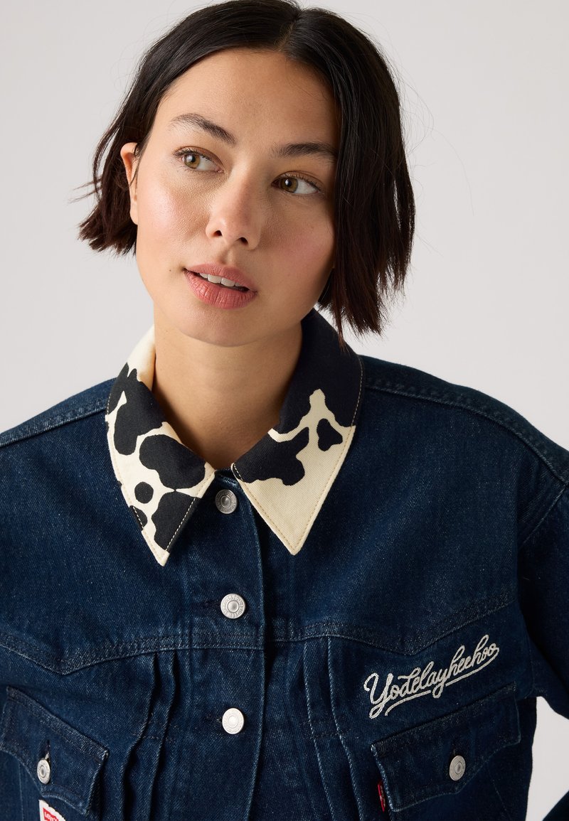 Giacca in denim di colore blu scuro, con colletto a motivo mucca color crema, bottoni a pressione argentati e dettaglio ricamato "Yodelayheehoo".