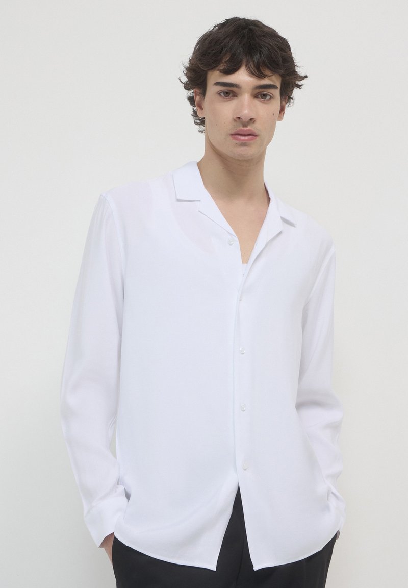 Jeune homme aux cheveux bruns portant une chemise blanche ample à boutons et un pantalon noir, se tenant devant un fond clair.