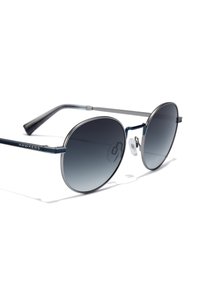 Ochelari de soare rotunzi cu lentile în degrade întunecat, ramă metalică de culoare albastră și suporturi pentru nas transparente. Numele mărcii "HAWKERS" gravat pe tâmplă.