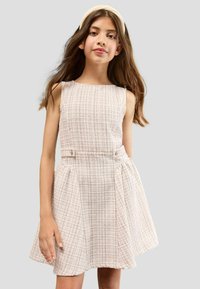 Ein Mädchen mit langen braunen Haaren trägt ein ärmelloses, hellrosa kariertes Kleid und ein passendes Stirnband und steht vor einem schlichten hellgrauen Hintergrund.