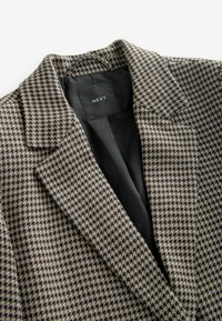 Houndstooth-mønstret blazer i beige og sort stof, med en klippet krave og sort foring. Mærket viser "NEXT."