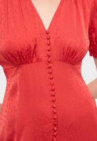 Acercamiento a un vestido rojo con lunares texturizados, botones cubiertos de tela en la parte delantera, escote en V y detalles fruncidos debajo del busto.