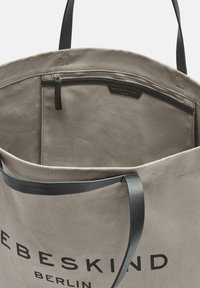 Beżowa torba materiałowa z czarnymi uchwytami, z wewnętrzną kieszenią na zamek błyskawiczny i napisem "LIEBESKIND BERLIN" na przedzie.