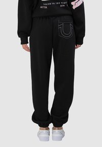 Pantalon de survêtement noir avec taille élastique, chevilles resserrées et une poche décorative avec couture à l'arrière. Tissu doux avec une finition texturée.