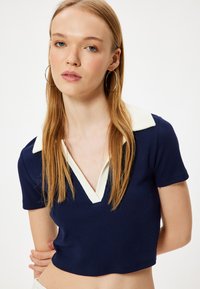 Top corto navy con scollatura a V profonda e colletto a coste bianche in contrasto. Le maniche corte e il tessuto testurizzato aggiungono dettaglio.