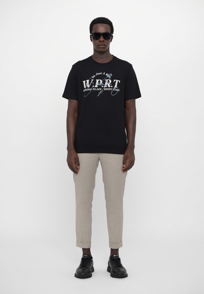 Tricou din bumbac negru cu text grafic "W.P.T." în alb și albastru, asortat cu pantaloni specifice de un bej deschis și adidași negri, stând pe un fundal alb.