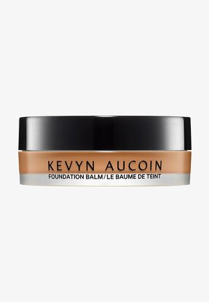 Baume de la Fondation Kevyn Aucoin dans un récipient rond transparent. Formule beige à l'intérieur, avec un couvercle noir mat et une bande argentée accentuée comportant le texte de la marque.