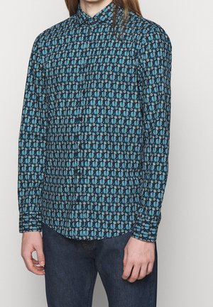 Homme portant une chemise boutonnée à manches longues avec un motif géométrique bleu, noir et blanc et un jean bleu foncé sur fond uni.
