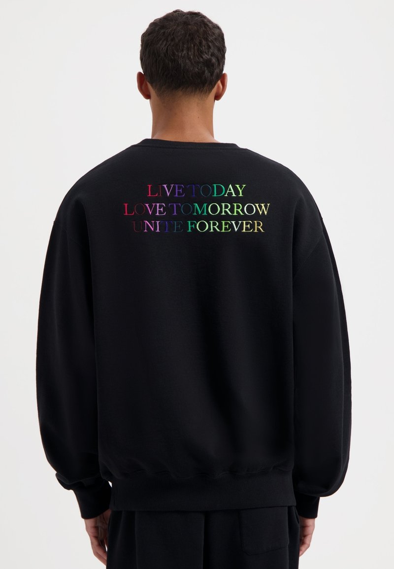 Zwarte sweatshirt met lange mouwen, een ronde hals en geribde manchetten; heeft op de achterkant een regenboogkleurige tekst: "LEEF VANDAAG, LOF MORGEN, VERENIG VOOR ALTIJD."