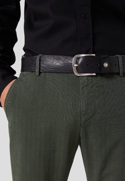 Cintura in pelle nera con una fibbia in metallo argentato, indossata in vita sopra pantaloni grigio scuro con una texture a spina di pesce.