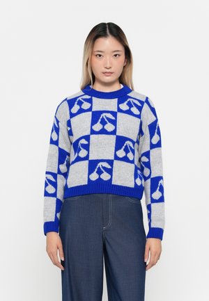 Jonge vrouw in een blauw-grijs geblokt truitje met witte kersenpatronen, gecombineerd met donkerblauwe wijde broek, staande tegen een witte achtergrond.
