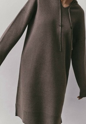 Braunes, langärmliges, knielanges Kapuzenkleid aus Strick mit Kordelzug, getragen von einer Person vor einem schlichten hellen Hintergrund, Arme leicht ausgestreckt.