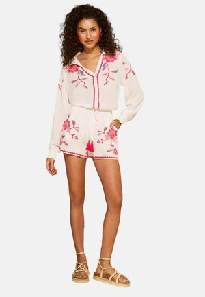 Femme portant une combinaison blanche à manches longues avec broderie florale rose, taille à cordon avec pompons et sandales beige en corde, debout avec une main dans la poche.