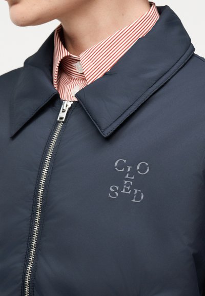 Άτομο που φοράει ένα ναυτικό μπουφάν με φερμουάρ πάνω από ένα κόκκινο και λευκό ριγωτό πουκάμισο με κουμπιά, με διασκορπισμένο κείμενο «ΚΛΕΙΣΤΟ» στο μπουφάν.
