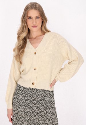 Crèmekleurige gebreide cardigan met een V-hals, drie bruine knopen en een geribde zoom. Draagt over een donkergroene rok met bloemenpatroon. Lichtgewicht textuur.