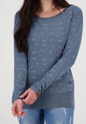 Femme portant un pull bleu à manches longues avec de petits motifs d'oiseaux, des détails de boutons près de l'encolure, et un jean bleu usé.
