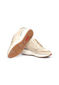 Zapatillas de cuero beige con suela de goma texturizada, que cuentan con un cierre de cordones en la parte delantera, perforaciones y un acento rojo en la suela.