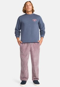 Dark blue sweatshirt with a round neckline and printed logo; paired with light pink corduroy pants; model wearing dark shoes.