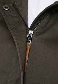 Chaqueta verde oliva con una cremallera visible, que cuenta con una pestaña de metal plateado y un acento de cuero marrón cerca de la cremallera. Tejido suave y texturizado.