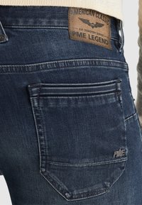Krupan plan tamnoplavih traperica s poleđinom džepa s izvezenim PME logotipom i smešnom kožnom etiketom na kojoj piše "American Classic PME Legend."