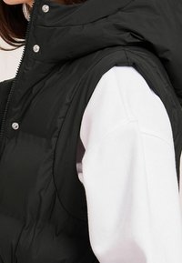 Czarna pikowana kamizelka z wysokim kołnierzem i zapięciem na zamek, z błyszczącymi zatrzaskami i gładką teksturą, noszona na białej bluzie.