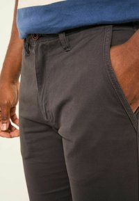 Dark gray cotton pants with a smooth texture, featuring a side pocket and a button closure at the waist, partially covered by a blue shirt.