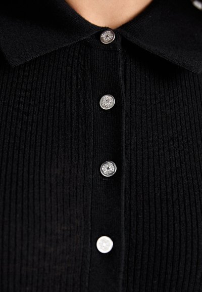 Haut en tricot noir à côtes avec col, comportant cinq boutons décoratifs argentés sur le devant. Texture douce avec un léger éclat.