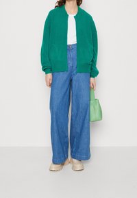 Grön stickad kofta, vit topp, blå vida jeans och beige skor. Håller en liten grön handväska. Enkelt mönster, inga synliga mönster.