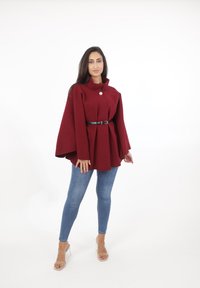 Capa de lana burdeos con un cuello alto, cierre de botones y un cinturón negro. Combinada con jeans ajustados y sandalias transparentes de tacón cuadrado.