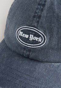 Sötétkék farmer baseball sapka fehér, kiemelt hímzett "New York" logóval, amely ovális alakú. Jellemzi a hajlított karima és a texturált anyag.