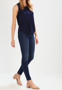 Granatowa bluzka bez rękawów z plisami z przodu, połączona z ciemnoniebieskimi dżinsami skinny i cielistymi butami na słupku z ostro zakończonym noskiem.