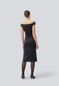 Svart off-shoulder kjole med en kroppsnær silhuett, knelengde og glatt stoff; kombinert med gjennomsiktige svarte strømpebukser og hælsandaler.