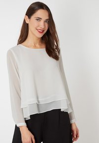 Blusa blanca de capas con mangas largas, fabricada en un tejido ligero y transparente, y con un escote redondeado. Llevada sobre pantalones oscuros.