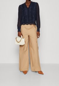 Marineblå spetsblus med långa ärmar och vågiga kanter, tillsammans med krämfärgade cargobyxor och orange spetsiga klackar, hållandes en liten vit väska.