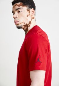 Piros pamut póló, kerek nyakkivágással és rövid ujjakkal, fekete Air Jordan logóval a bal ujján. Simaság jellemzi.