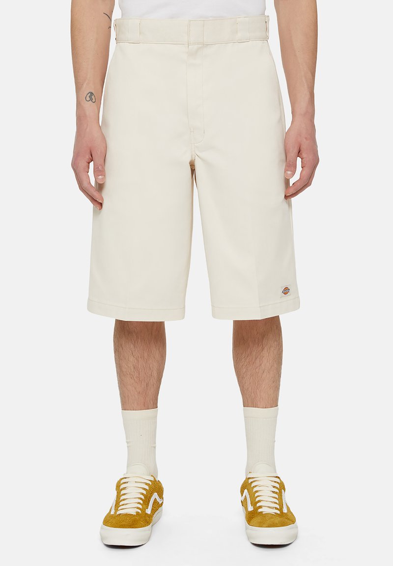 Shorts en coton beige avec une coupe droite, un devant plat et des poches latérales. Présente un petit patch logo près de l'ourlet. Portés avec des baskets jaunes.