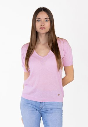 Vrouw met lang, steil bruin haar, gekleed in een lichtroze, korte mouw V-hals trui en lichtblauwe spijkerbroek, tegen een witte achtergrond.