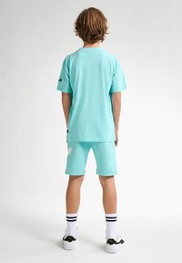 Conjunto de camiseta y pantalones cortos en color turquesa, material de algodón, mangas cortas, corte relajado, los pantalones cortos presentan un parche blanco como acento, combinados con zapatillas blancas y negras.