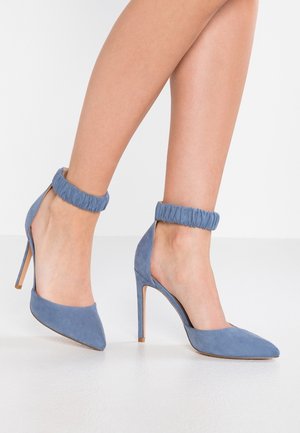 Nogi w niebieskich zamszowych szpilkach z ostrymi noskami i marszczonymi paskami wokół kostek na białej podłodze.