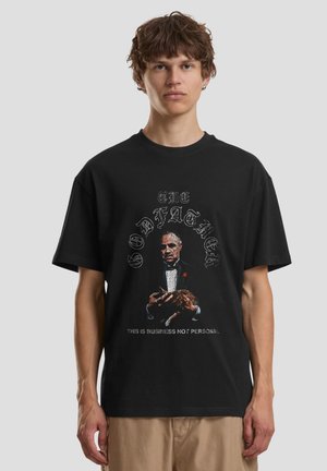 Młody mężczyzna w czarnej koszulce z grafiką starszego mężczyzny w smokingu trzymającego kota, z tekstem nad i pod obrazkiem.