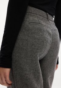 Calças de cintura alta em padrão cinza de espinha de peixe, com bolsos frontais, corte ajustado e textura suave. Top preto de mangas longas visível.