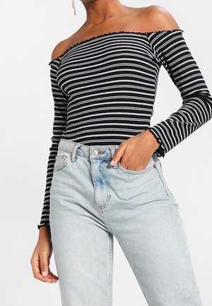 Frau trägt ein schwarz-weiß horizontal gestreiftes, schulterfreies Langarmoberteil mit hellblauen hochgeschnittenen Jeans, die Hand in der Tasche.