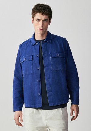 Young man with short brown hair wears a blue button-up jacket over a black shirt and light-colored pants, standing against a plain background.