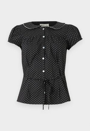 Blouse noire à manches courtes avec pois blancs, col arrondi bordé de blanc, boutons à l'avant et lien de serrage à la taille.