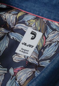 Denim stof met een binnenvoering met bloemafdrukken in blauw, roze en geel. Heeft een wit J&JOY label met zwarte tekst, waarin staat "Ontworpen in België."