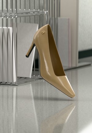 Beež värvi terava ninaga kõrge kontsaga king kuldse väikese embleemiga, püsti asetatud peegeldavale pinnale metallist failikorraldaja lähedal.