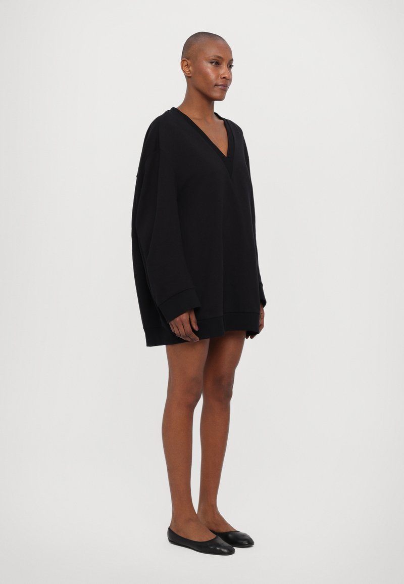Robe hoodie oversize noire avec un décolleté en V profond, des manches longues et des poignets côtelés. Texture de tissu lisse et doux. Ballerines noires plates.