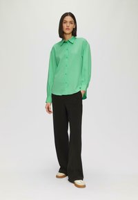Camicia verde con bottoni, finitura testurizzata, maniche lunghe e colletto, abbinata a pantaloni neri a gamba larga e sneakers beige.